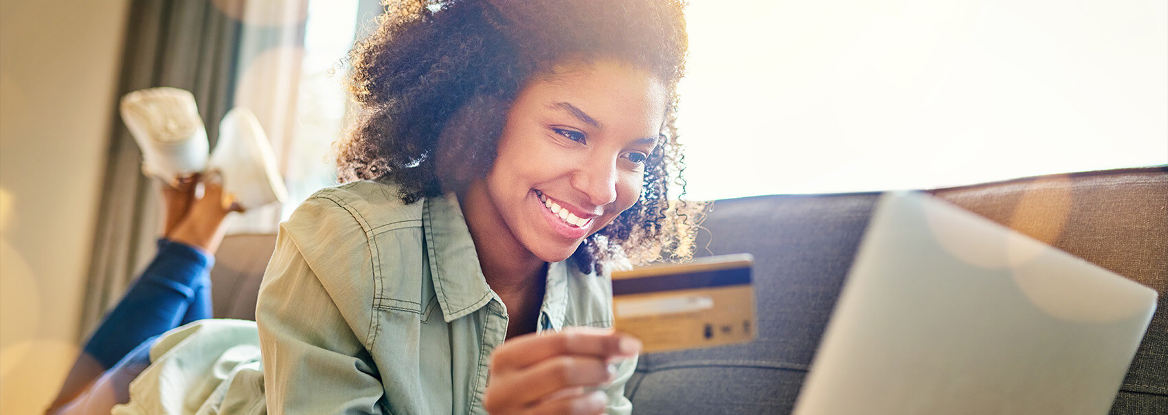 young adult woman laying on stomach on couch with her laptop, holding a credit card in one hand