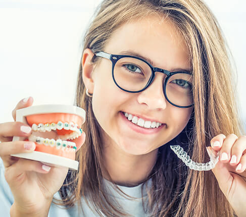 teenage girl holding up a set of clear aligners and model of teeth with braces attached